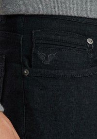 Jeans de denim negro para hombre con una textura de espiga, que cuentan con bolsillos delanteros y traseros, un botón de metal y un detalle de logo bordado.
