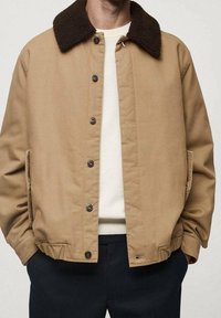 Beżowa kurtka z brązowym, puszystym kołnierzem, z guzikami z przodu, bocznymi kieszeniami i luźnym krojem. Nosi się ją na białym swetrze.