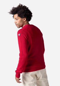 Rotem Strickpullover mit rundem Ausschnitt, gerippten Bündchen und seitlichen Streifen. Inklusive eines kleinen Logo-Patches an der linken Schulter.