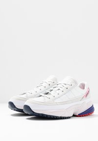 Vita idrottsskor med överdel i mocka och mesh, med snörning, flerfärgad sula med lila, rosa och svarta detaljer.