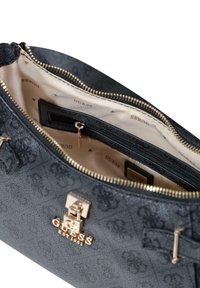 Bolso negro texturizado con herrajes dorados, cierre de cremallera y placa con el logotipo. El interior presenta tela crema con un patrón repetido del nombre de la marca.