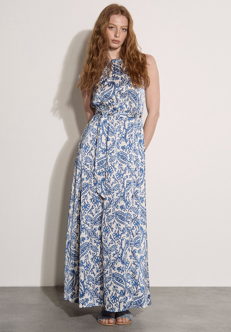 Femme aux longs cheveux roux portant une robe maxi blanche sans manches avec des motifs floraux bleus, debout devant un fond clair uni.