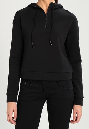 Osoba nosząca czarną bluzę z kapturem, z przednim zamkiem i sznurkami, w połączeniu z czarnymi jeansami, stojąca na neutralnym tle.