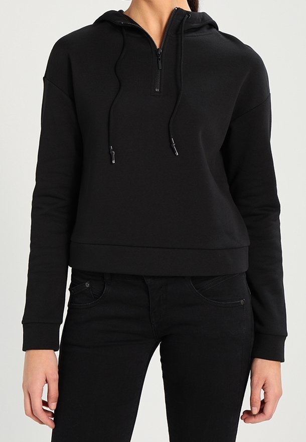Personne portant un sweat à capuche noir avec une fermeture éclair avant et des cordons, associée à un jean noir, debout devant un fond neutre.