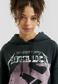 Czarna bluza z kapturem z przodu z nadrukiem graficznym zawierającym napis "Metallica". Materiał wygląda na miękki; sznurki z metalowymi końcówkami akcentują kaptur.