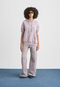 Ανοιχτό μωβ φούτερ με κουκούλα, λογότυπο, τσέπη καγκουρό και κορδόνια· συνδυάζεται με αντίστοιχο παντελόνι με φαρδιά μπατζάκια και μαύρα παπούτσια πάνω σε μπλε χαλί.