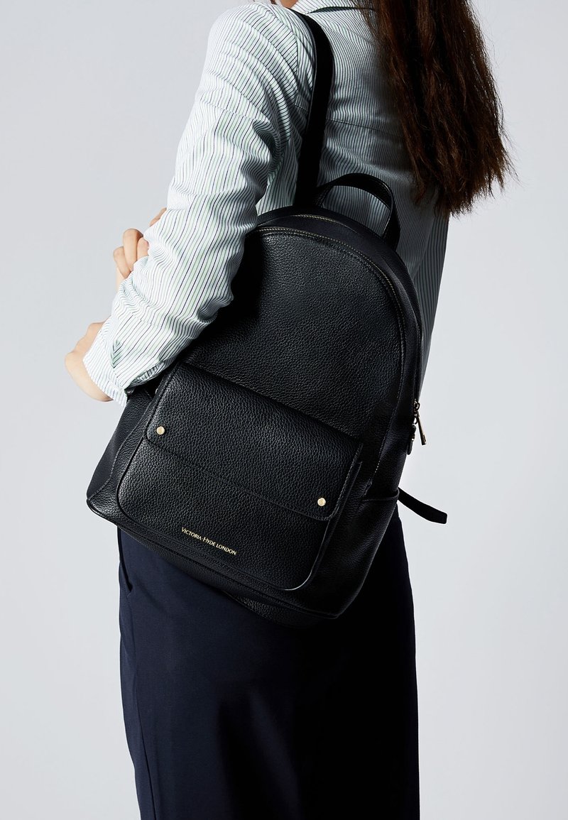 Femme portant une chemise rayée et une jupe foncée, portant un sac à dos en cuir noir texturé avec une poche avant et des clous dorés.