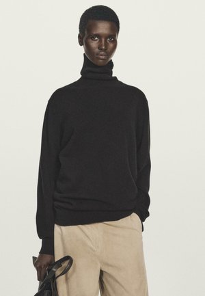 Person in einem schwarzen Rollkragenpullover und beigen Hosen, der eine schwarze Handtasche hält, vor einem hellen, einfarbigen Hintergrund.