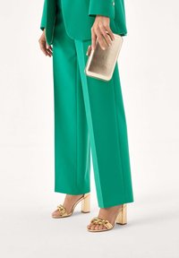 Grøn skræddersyet jakkesæt med vide ben, kombineret med gyldne højhælede sandaler med kædedetalje og en gylden struktureret clutch.