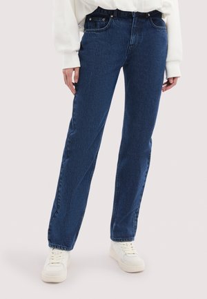 Person iført mørkeblå jeans med lige ben, en hvid langærmet skjorte stoppet i bukserne og hvide sneakers, stående på en lys baggrund.