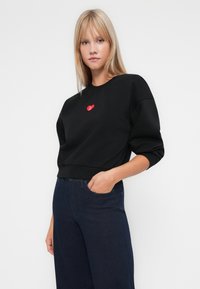 Hanorac negru cu un patch în formă de inimă roșie, având decolteu rotund, croială scurtă și o croială lejeră. Purtați cu blugi bleu închis.