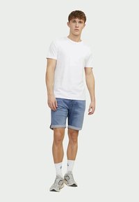 Young man standing, wearing a plain white T-shirt, rolled-up blue denim shorts, white crew socks, and gray sneakers, against a plain white background.