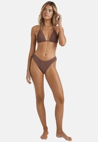 Bikini triangolare marrone con tessuto texturizzato, caratterizzato da un design minimalista e una parte inferiore alta. La modella è in piedi a piedi nudi con una mano tra i capelli.