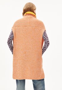 Oranžová pletená vesta s texturovanými vzory, vysokým límcem a otevřenými rukávy, vrstvená přes košili s dlouhým rukávem s vzorem.