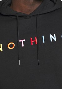 Sudadera negra con la palabra "NOTHING" bordada en letras multicolores. Presenta cordones con puntas negras y un tejido suave y texturizado.