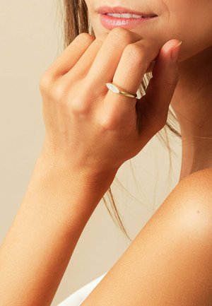 Goldring mit ovaler, strukturiertem Kopf, getragen an einer Hand mit einem natürlichen Hautton. Der Hintergrund ist neutral und sanft beleuchtet.