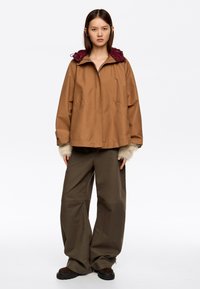 Brązowa kurtka z kapturem, z bordowym sznurkiem, kieszeniami po bokach i lekkimi akcentami sztucznego futra. Połączona z luźnymi oliwkowymi spodniami.