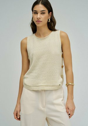 Jeune femme portant un top en maille beige sans manches avec des boutons sur le côté, un pantalon couleur crème, des boucles d'oreilles en or et un bracelet en or, debout devant un fond gris.