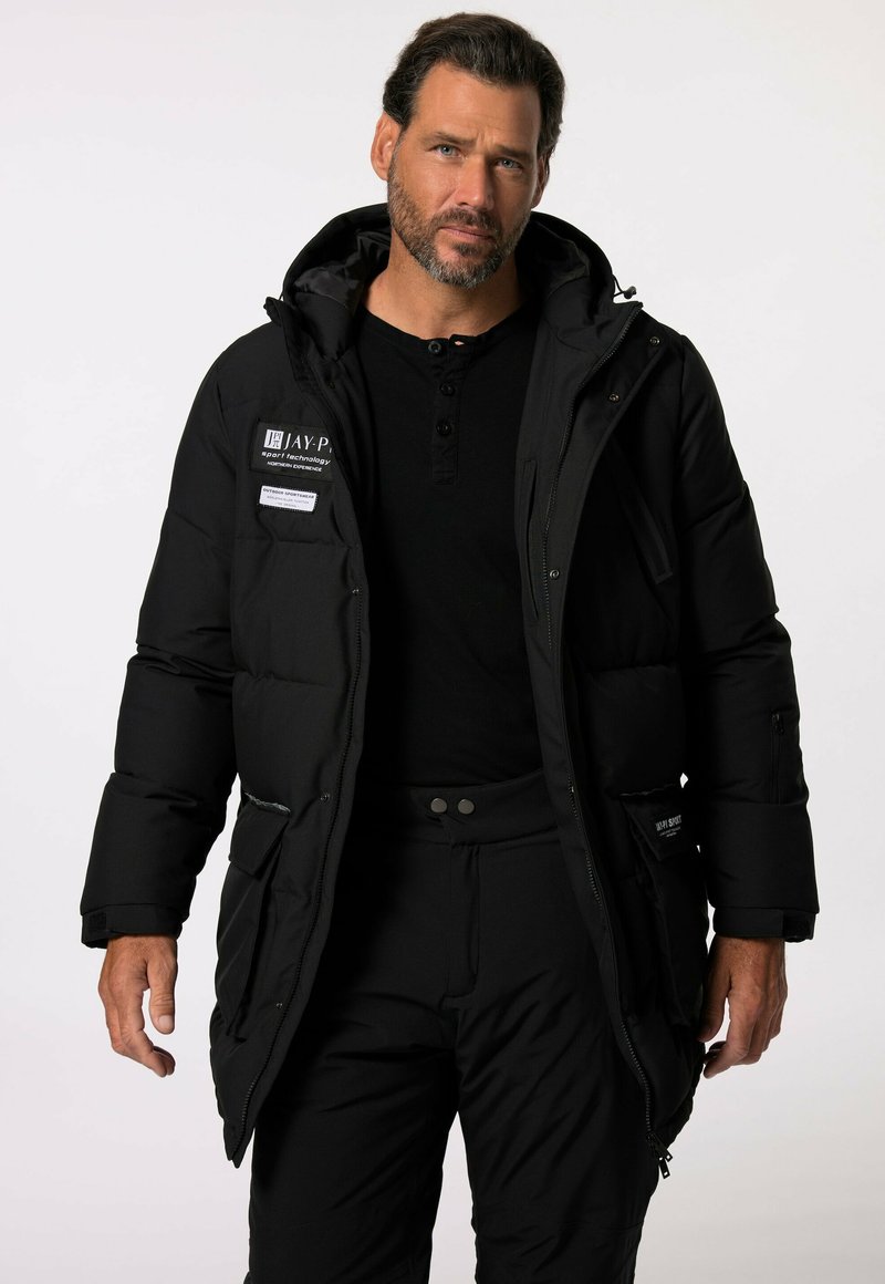 Parka nera imbottita con colletto alto, numerose tasche e chiusura con zip. Indossata sopra una maglietta henley nera, caratterizzata da una trama robusta.