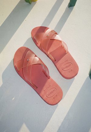 Paire de sandales rouges Havaianas avec des brides croisées reposant sur une surface blanche avec des ombres vertes.