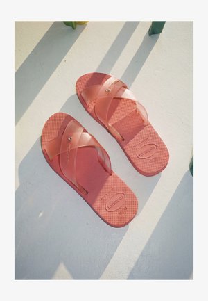 Een paar rode Havaianas sandalen met gekruiste bandjes, liggend op een wit oppervlak met groene schaduwen.