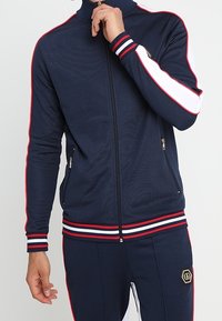 Chaqueta de chándal de cremallera en color marino con cuello alto, franjas laterales blancas y rojas, y puños acanalados. Presenta bolsillos con cremallera y material texturizado.