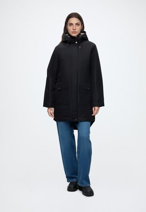 Femme portant un manteau d'hiver noir à capuche, un jean bleu et des chaussures noires, debout devant un fond blanc uni.