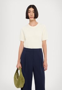 Top crème à manches courtes à côtes associé à un pantalon taille haute bleu marine foncé. Le mannequin tient un sac à main en daim vert olive doux.