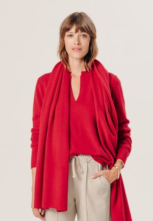 Pull rouge en tricot avec un décolleté en V profond, superposé avec une écharpe oversized assortie. Pantalons beiges à cordon de serrage et un bracelet en or visible.