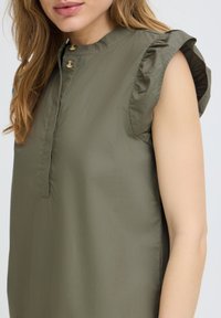 Femme portant un chemisier sans manches vert olive avec des volants aux épaules et un col haut boutonné sur fond blanc.