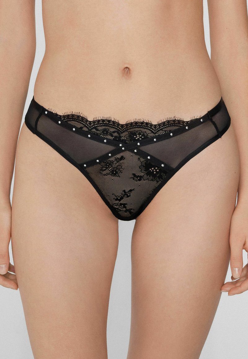 Tanga de encaje negro con un ribete de encaje en forma de pico, patrón floral y pequeñas perlas decorativas a lo largo de la cintura y los laterales. Material de malla transparente.