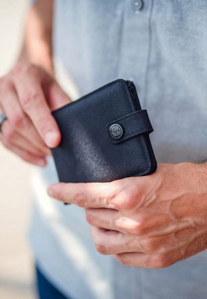 Black fabric wallet with a button closure, featuring a textured surface and a small, circular logo on the front. Held in a hand.