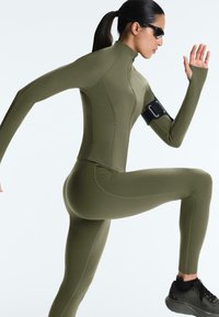 Femme en tenue de sport vert olive et lunettes de soleil levant un genou et un bras, portant un brassard noir et des baskets noires, en train de faire du jogging ou de l'exercice.