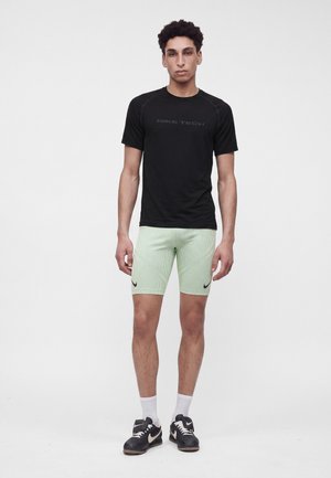 Un jeune homme se tient debout, portant un t-shirt Nike Tech noir, un short côtelé vert clair avec des logos Nike, des chaussettes blanches et des baskets Nike noires.