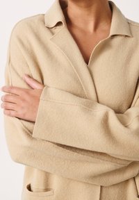 Beige wollen jasje met een ingesneden kraag, lange mouwen en een voorzak. De stof lijkt zacht en textuur te hebben met zichtbare naden.