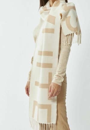 Femme portant un haut à manches longues beige et un pantalon, avec une écharpe à motif géométrique blanc et beige drapée sur les épaules.