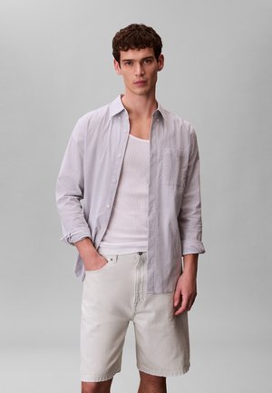 Unge mand iført hvid tanktop, lyse, stribede, knappede skjorte med ærmerne rullet op og hvide shorts til knæene, stående mod en ensfarvet grå baggrund.