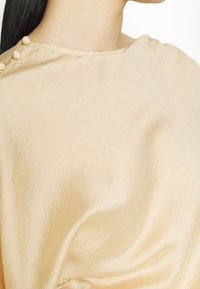 Beige kangas, ambapo kuna ngozi laini, neckline ya boat, na mapambo ya button kwenye mshono wa bega.