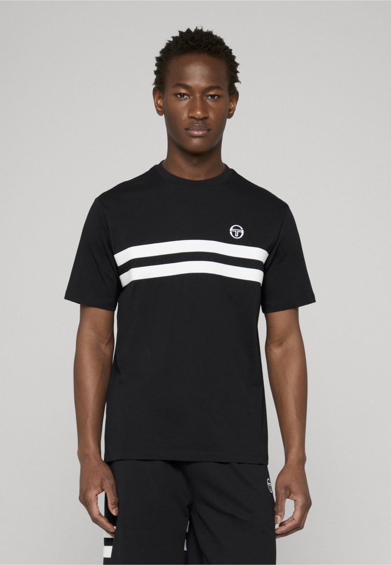 Mann iført svart kortermet skjorte med to hvite horisontale striper og en liten sirkulær logo på brystet, kombinert med svarte shorts.