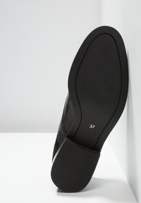 Chaussure en cuir noire vue du côté de la semelle, taille 37 imprimée sur la semelle, positionnée contre un mur d’angle blanc.