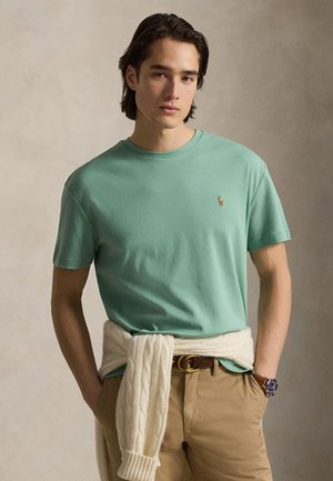 Νεαρός άνδρας με μεσαίου μήκους σκουρόχρωμα μαλλιά, που φοράει ένα ανοιχτό πράσινο μπλουζάκι, μπεζ παντελόνι και ένα κρεμ πουλόβερ δεμένο γύρω από τη μέση του.