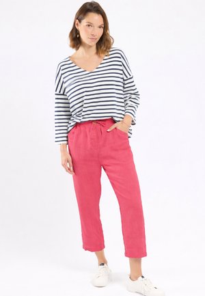 Femme debout portant un haut à manches longues blanc à rayures bleu marine, un pantalon court rouge et des baskets blanches, posant avec une main dans la poche.