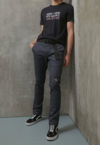 Czarna koszulka z grafiką "New Balance Invitational", połączona z ciemnoszarymi spodniami z paskiem i logo, uzupełniona czarno-białymi trampkami.