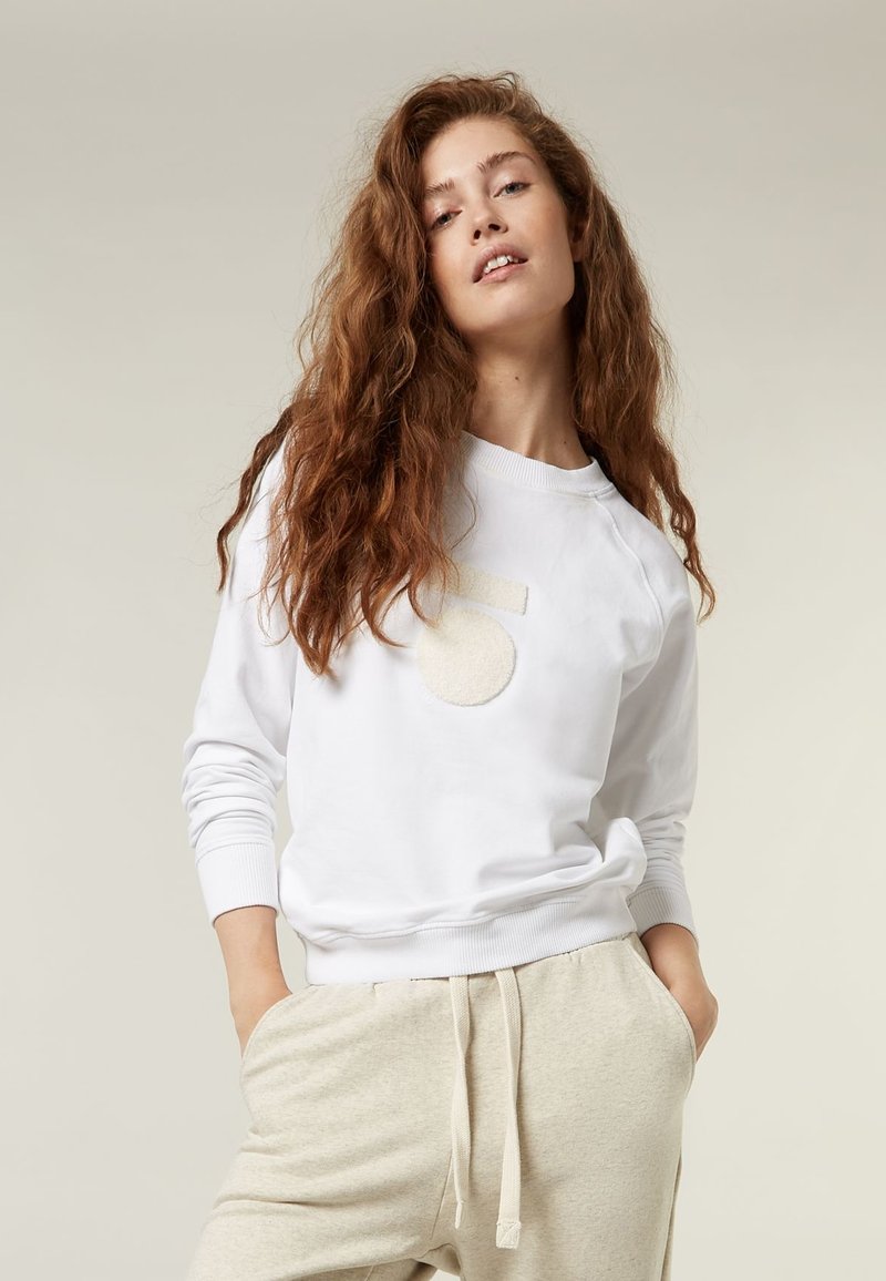 Felpa bianca con un'appliqué circolare testurizzata e vestibilità oversize, abbinata a joggers beige chiaro. Il tessuto sembra morbido e informale.