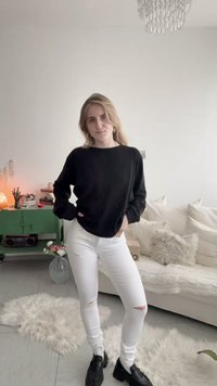 Γυναίκα με ξανθά μαλλιά που φοράει μαύρο μπλουζάκι μακρυμάνικο, λευκό σκισμένο τζιν και μαύρα παπούτσια, στέκεται σε ένα φωτεινό σαλόνι με καναπέ, χαλί και πράσινη ντουλάπα.