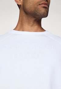 Hvit hettegenser med preget "TED BAKER SPORT"-tekst foran, med rund hals og raglanermer. Glatt stofftekstur.