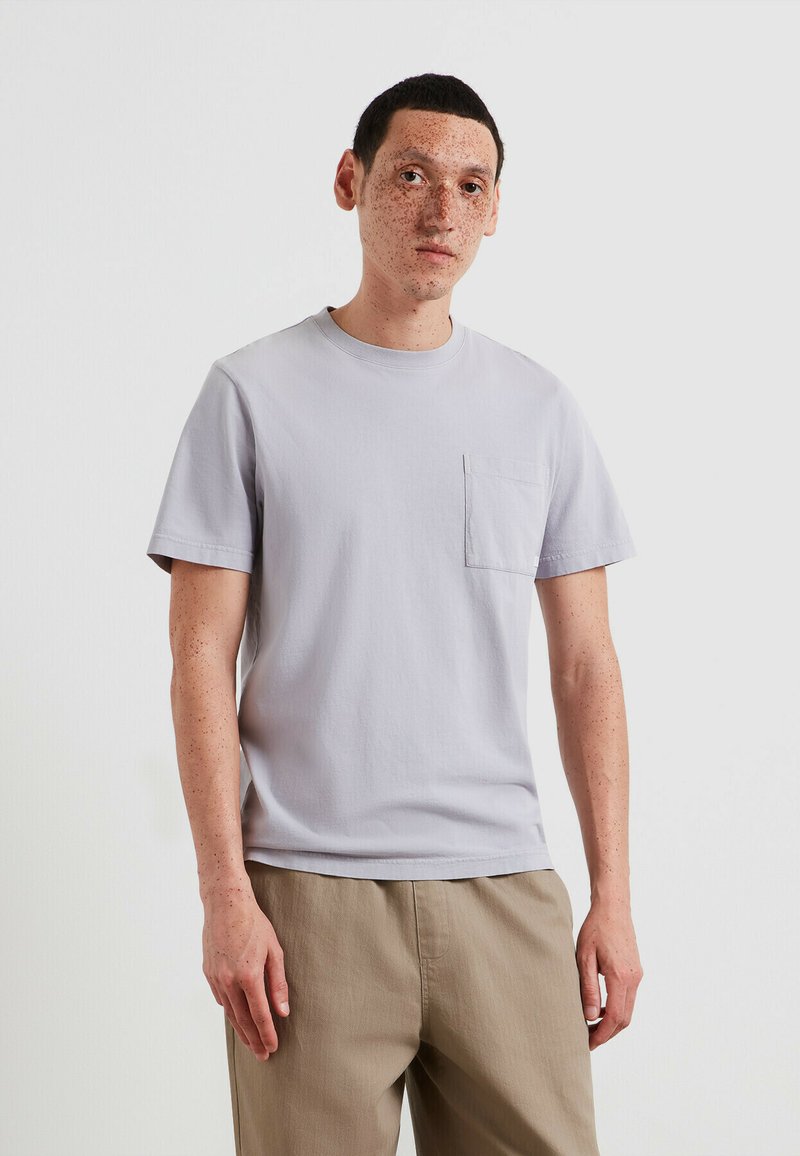 T-shirt en coton gris clair avec col rond, manches courtes et une poche poitrine, porté avec un pantalon beige. Texture lisse, design simple.