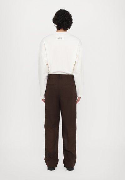 Back view of a model wearing a long-sleeve off-white shirt and loose brown trousers with a subtle texture and rear pockets.
