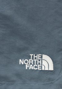 Λευκό κεντημένο λογότυπο The North Face σε μπλε-γκρι υφασμάτινη υφή με ορατή ραφή κοντά στην άκρη.