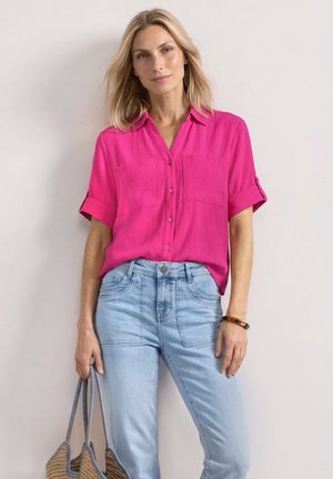 Femme blonde portant un chemisier rose vif à manches courtes et un jean bleu clair tenant un sac fourre-tout tissé avec des poignées grises sur un fond uni.